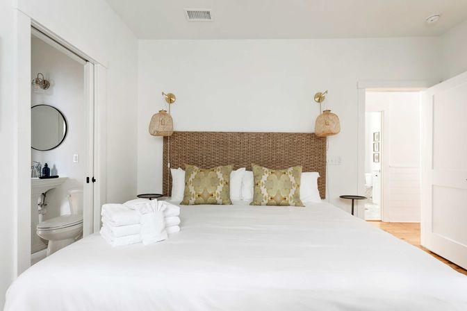 Bright, minimalist coastal-inspired bedroom with white linens, woven rattan headboard, green patterned throw pillows, hanging wicker sconces, en-suite bathroom visible and neatly folded white towels on the bed.
