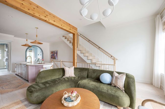 Bright open-plan coastal-style living room and kitchen with a curvy olive-green sectional, round wood coffee table, light wood beam and staircase, pastel island and rattan pendant lights.