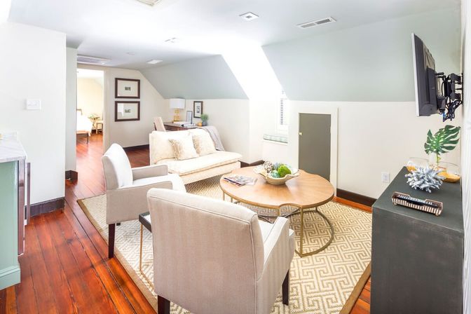 Bright cozy attic living room with sloped ceiling and skylight, hardwood floors, neutral sofa and two armchairs arranged around a round wooden coffee table on a patterned rug, wall-mounted TV and styled console.