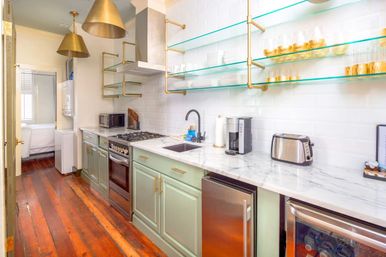 Bright, stylish galley apartment kitchen with sage-green lower cabinets, marble countertop, white subway-tile backsplash, open glass shelves with brass brackets, brass pendant lights, stainless-steel range and appliances, black faucet and warm hardwood floors.