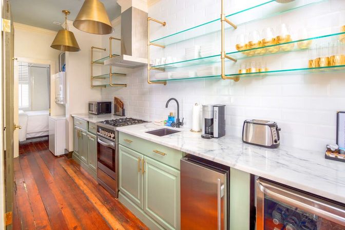 Bright, stylish galley apartment kitchen with sage-green lower cabinets, marble countertop, white subway-tile backsplash, open glass shelves with brass brackets, brass pendant lights, stainless-steel range and appliances, black faucet and warm hardwood floors.