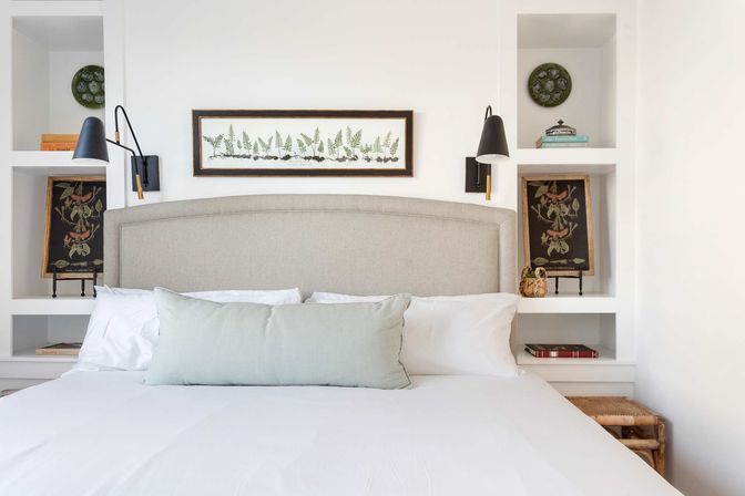 Bright, serene coastal-inspired bedroom with a beige upholstered headboard, white built-in shelves, framed fern print, black swing-arm wall sconces, botanical decor and a pale green lumbar pillow on crisp white linens.