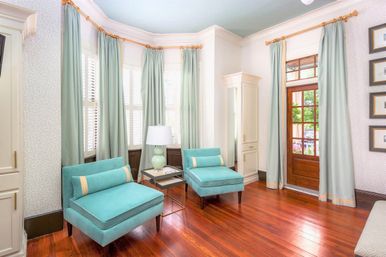 Sunlit bay-window living room with two turquoise lounge chairs, pale green floor-length curtains, glossy hardwood floors, glass-pane wooden door and a bedside table with lamp.