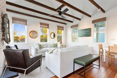 Bright coastal-style living room with vaulted ceiling and exposed dark beams, white shiplap walls, large windows with woven blinds, two white sofas with green pillows, a dark leather armchair, green bench on warm wood floors, round mirrors and a wall sign, and a small dining nook with wicker chairs.