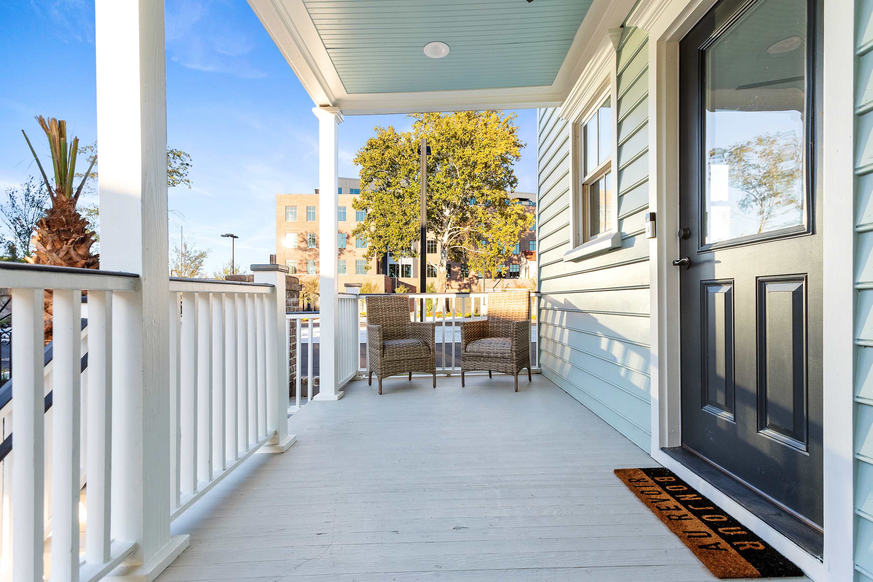 Sunny covered front porch with light-blue siding and black door, white railings, two wicker chairs and a doormat overlooking a tree-lined urban street and office building.