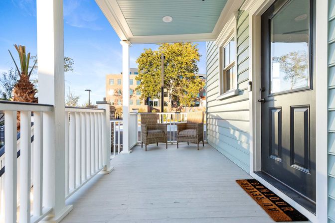 Sunny covered front porch with light-blue siding and black door, white railings, two wicker chairs and a doormat overlooking a tree-lined urban street and office building.