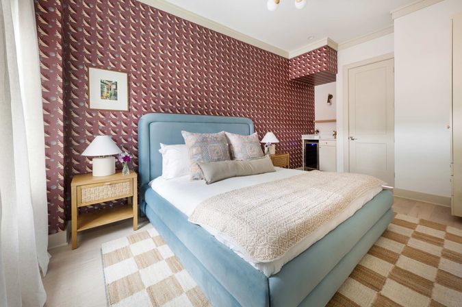 Stylish bright bedroom in a modern apartment with a light blue upholstered bed, patterned maroon wallpaper, woven nightstands with white lamps, decorative pillows and beige throw, checkerboard rug, and a small kitchenette with mini fridge.
