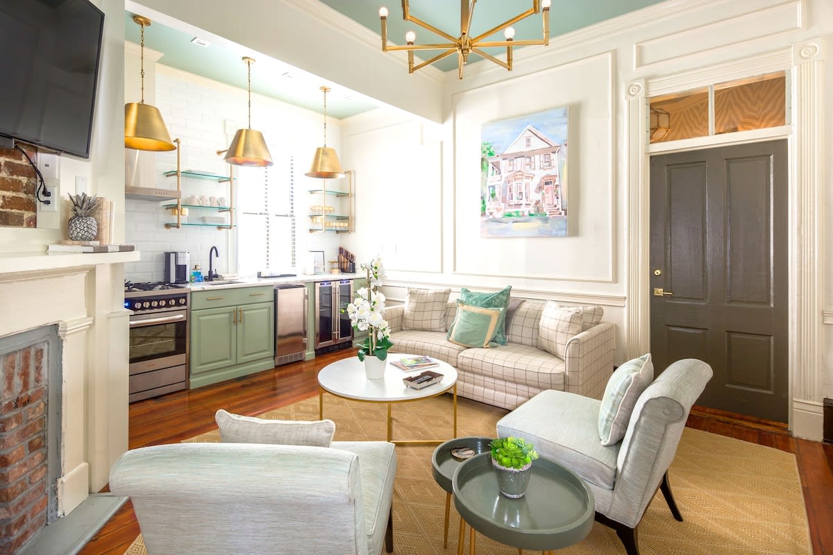 Sunlit open-plan living room and kitchen in a renovated townhouse — mint-green cabinets, brass pendant lights, plaid sofa with green accent pillows, round white coffee table, hardwood floors and a dark entry door.