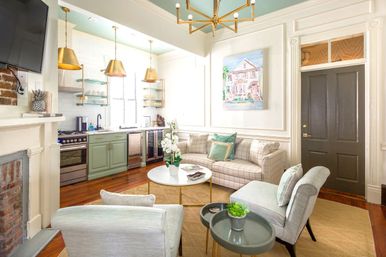 Sunlit open-plan living room and kitchen in a renovated townhouse — mint-green cabinets, brass pendant lights, plaid sofa with green accent pillows, round white coffee table, hardwood floors and a dark entry door.