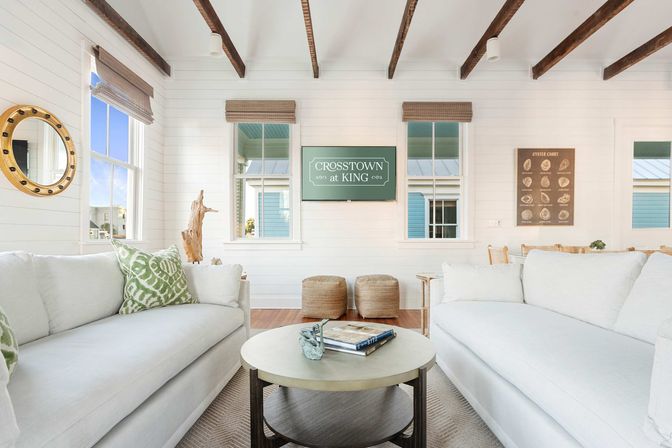 Bright coastal living room with white shiplap walls and exposed dark wood beams, two white sofas facing a round coffee table with books and a seashell, woven ottomans, bamboo shades on windows and nautical decor.