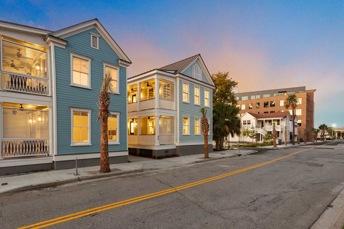 Charming sunset coastal street with two-story pastel blue homes, screened porches and warm lit windows, small palm trees lining a quiet road and a brick mid-rise in the background.