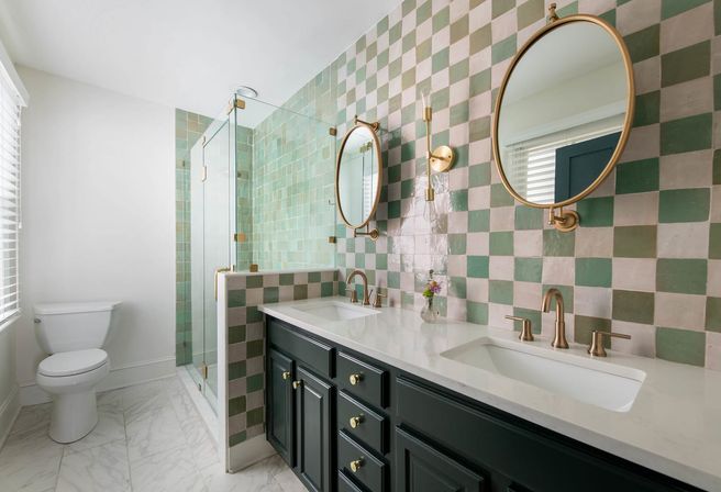 Bright modern master bathroom with green-and-white checkered tile wall, glass shower, double white quartz sinks, brass faucets and round mirrors on a marble-look floor.