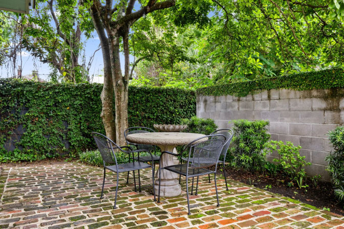 Shaded backyard garden patio with mossy brick pavers, stone pedestal table and four black metal chairs beneath leafy trees, ivy-covered wall and small shrubs.
