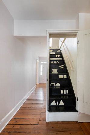 Bright narrow hallway with polished hardwood floors and a quirky black staircase painted with white geometric shapes and a simple wall handrail.