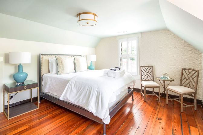 Cozy bright attic bedroom with white linens on a metal bed frame, teal bedside lamps on dark wood nightstands, warm polished hardwood floors, pale blue sloped ceiling, shuttered window letting in natural light, and two woven-back chairs with a small glass side table.