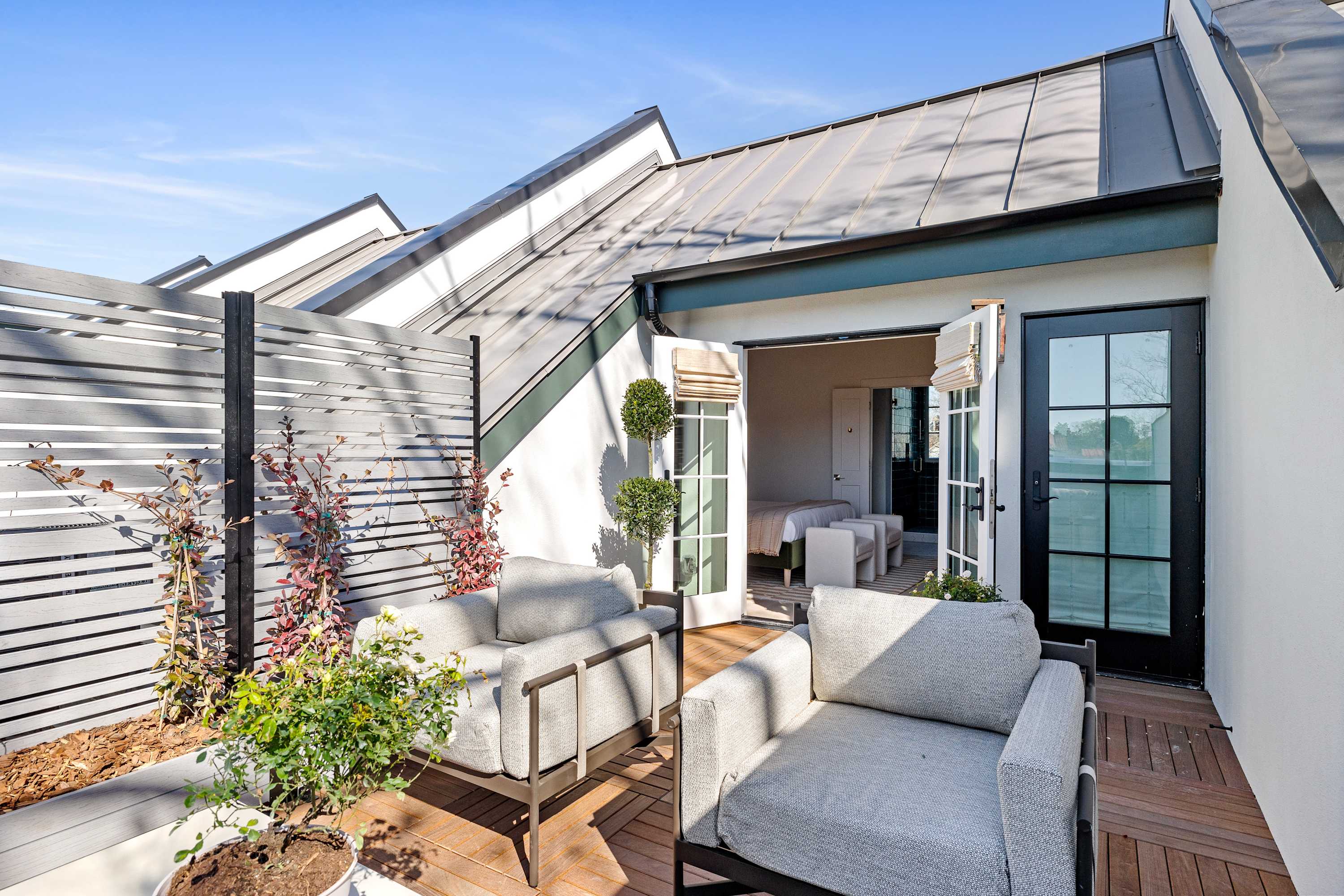 Sunlit modern rooftop terrace on an urban loft with cozy gray lounge chairs, wooden deck, potted plants, slatted privacy screen and French doors opening to a bedroom.