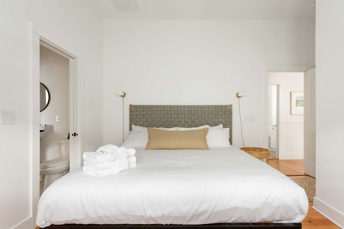 Bright minimalist bedroom with hardwood floor, king bed with woven gray headboard, crisp white linens and beige lumbar pillow, folded white towels on the bed, brass wall sconces and open doorways to adjoining bathrooms.