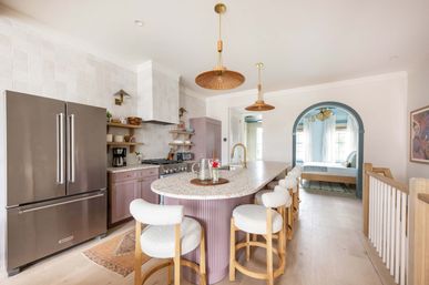 Bright modern kitchen with terrazzo island and mauve shaker cabinets, boucle bar stools, stainless French-door fridge, rattan pendant lights, arched blue doorway and light oak floors