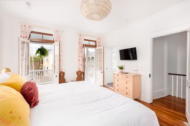 Bright airy bedroom with crisp white linens, yellow and red accent pillows, hardwood floors, light wood dresser and wall TV, French doors opening to a small balcony with a hanging fern and neighborhood views.