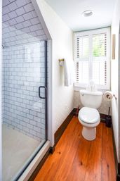Cozy bright bathroom with glass-door subway-tile shower, white toilet under shuttered window, warm hardwood floor and brass towel hook.