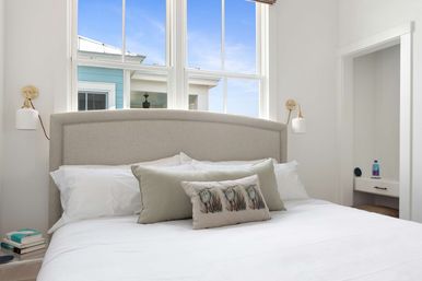 Bright coastal-style bedroom with neutral upholstered headboard, crisp white linens, decorative seagrass pillow, brass wall sconces, bedside books, and a large window with blue sky.