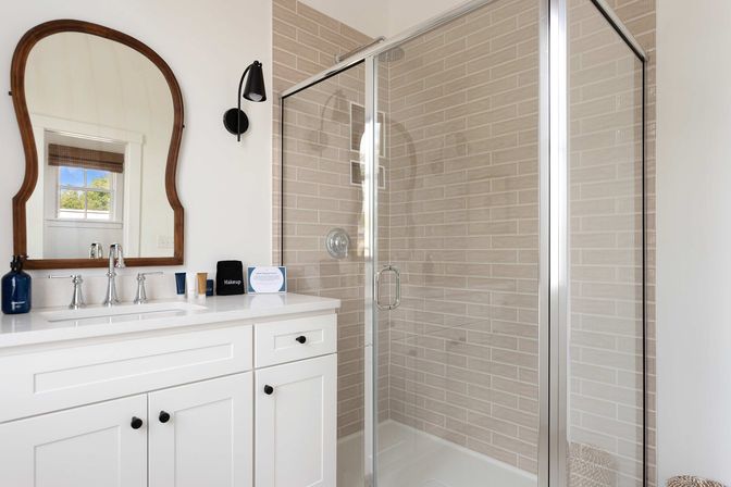 Bright modern bathroom with white vanity and quartz countertop, wood-framed mirror, chrome faucet, and glass-enclosed shower with beige subway tile and black accents.