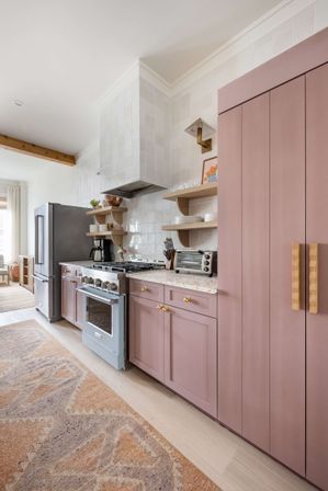 Sunlit modern kitchen interior with matte dusty-pink shaker cabinets, brass knobs and carved wood handles, stainless steel refrigerator, pale blue gas range, open wood shelves, white square tile backsplash, terrazzo-style countertop and patterned area rug.