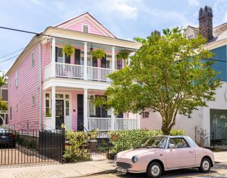Sunlit pastel pink two-story house with white double-deck porch and hanging ferns, a leafy front tree and a matching pink vintage convertible parked curbside on a tree-lined residential street.
