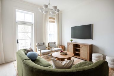 Sunlit contemporary living room with a curved olive-green sofa, round wooden coffee table, two light-upholstered armchairs, rattan-front media console and wall-mounted TV beneath tall windows with sheer curtains.