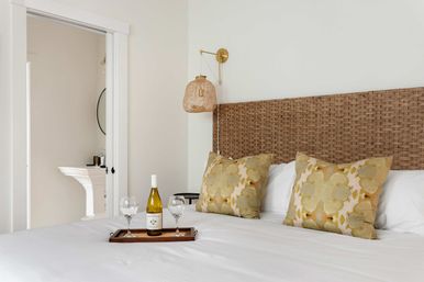 Bright coastal-style bedroom with a woven rattan headboard, two yellow floral throw pillows on crisp white bedding, a wooden tray holding a bottle of white wine and two glasses, and an open doorway revealing a pedestal sink and round mirror.