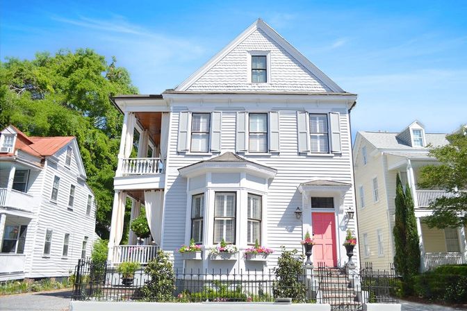 White Victorian-style house with white siding, bay window, second-story porch, pink front door, flower boxes and black iron fence under a bright blue sky
