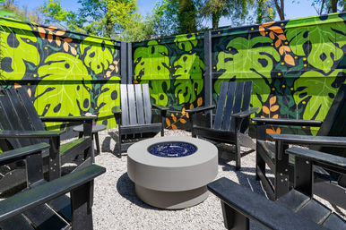 Outdoor private courtyard with black Adirondack chairs around a round modern fire pit on a gravel patio, bordered by a vibrant green tropical-leaf mural painted on a wooden fence.