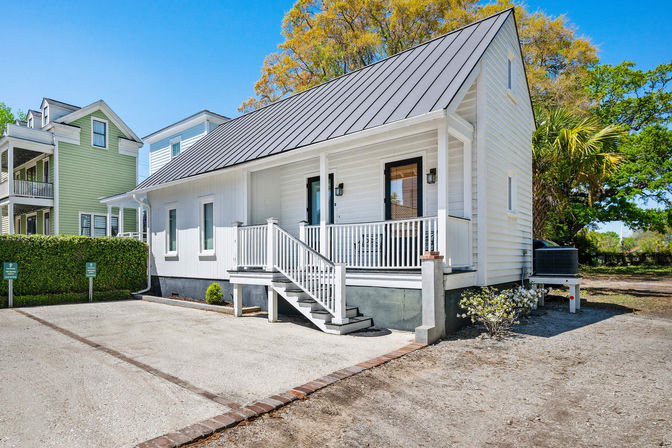 Bright white coastal cottage with black metal gable roof, front porch and steps, gravel parking area, palm tree and neighboring colorful houses under a clear blue sky
