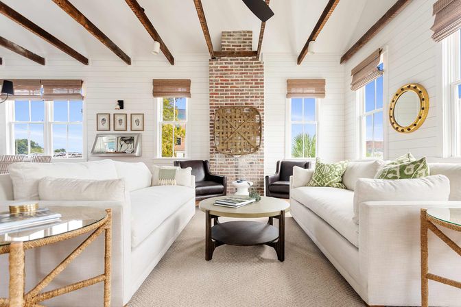 Sunlit coastal living room with vaulted ceiling and dark exposed beams, brick chimney, paired white sofas facing a round coffee table, wicker blinds and a nautical porthole mirror.