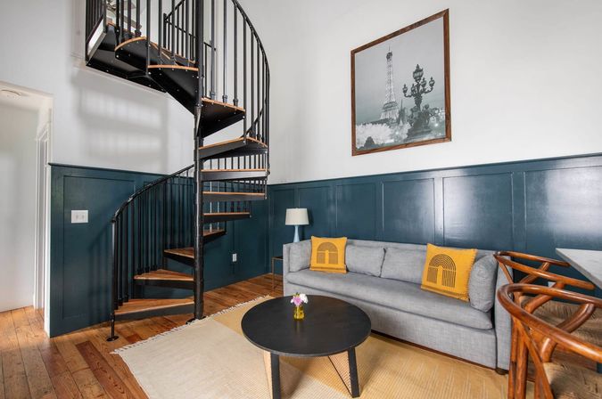 Cozy urban living room with black metal spiral staircase, deep teal wainscoting, gray sofa with yellow accent pillows, round black coffee table and framed Eiffel Tower wall art.