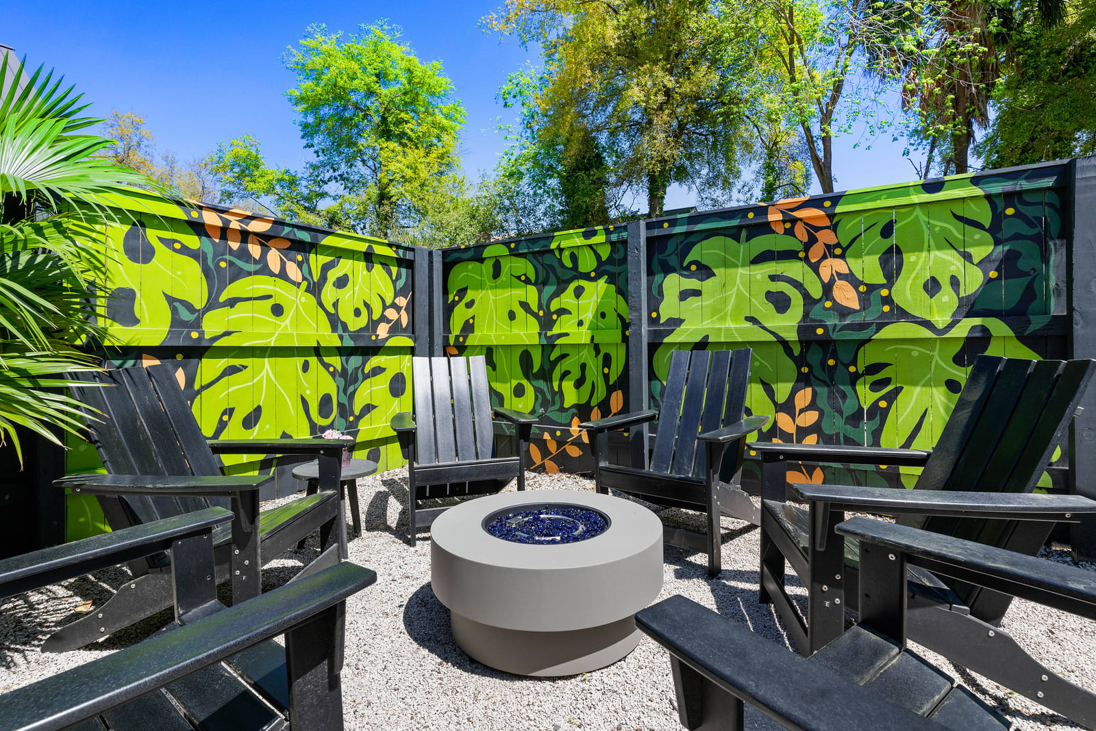 Sunny outdoor courtyard with black Adirondack chairs arranged around a modern round fire pit on gravel, set against a vibrant tropical-leaf mural painted on a privacy fence and surrounded by palm fronds and trees.