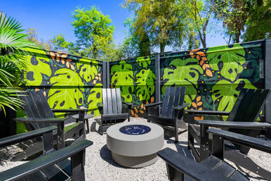 Sunny outdoor courtyard with black Adirondack chairs arranged around a modern round fire pit on gravel, set against a vibrant tropical-leaf mural painted on a privacy fence and surrounded by palm fronds and trees.
