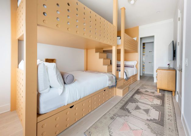 Bright modern bunk bedroom with peach-colored built-in twin bunks, stair access and under-bed drawers, patterned runner rug and wall-mounted TV — kid-friendly vacation rental room