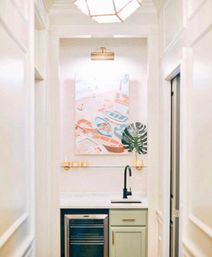 Bright hallway wet bar nook with pale green cabinet, mini fridge, marble countertop, matte-black gooseneck faucet, glass shelf, gold accents, herringbone wallpaper and cheerful pastel boat artwork.