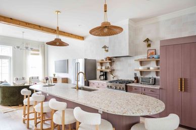 Sunlit open-plan kitchen with large terrazzo island, brass faucet and undermount sink, mauve cabinetry and hidden fridge, white boucle bar stools, rattan pendant lights, stainless range and a cozy living area visible in the background.