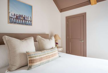 Cozy coastal-inspired bedroom with white bedding, oversized linen pillows and a striped lumbar cushion, round nightstand with wicker lamp and flowers, beach photo above the headboard, muted blush door and exposed wood beam.