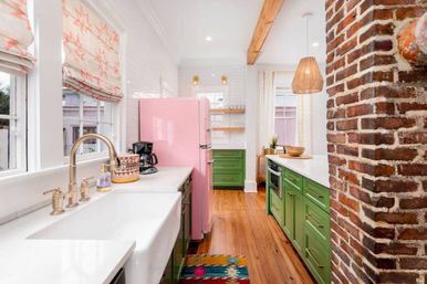 Sunny modern farmhouse kitchen with pink retro refrigerator, green shaker cabinets, white countertops, brass faucet, farmhouse sink, exposed brick column, wood floors and wicker pendant light.