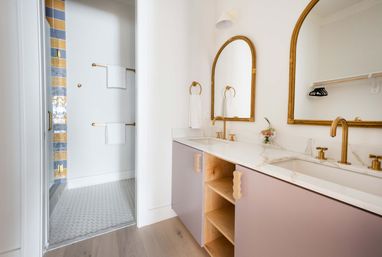 Bright modern bathroom with double marble vanity, blush cabinets, arched gold-framed mirrors, brass faucets, and adjacent walk-in shower with blue-and-gold accent tiles and hexagon floor tile.