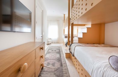 Bright airy modern bedroom in a family-friendly vacation rental with built-in wooden bunk beds featuring circular cutouts, cozy lower bunks, patterned runner rug, wooden dresser with round knobs, wall-mounted TV and sunlit window with sheer curtains.