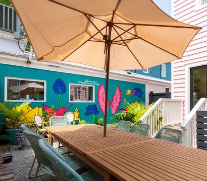 Vibrant courtyard patio with a long wooden dining table, large beige umbrella and woven chairs in front of a bright turquoise mural of flowers, birds and pink wings, flanked by pastel pink siding and potted tropical plants.