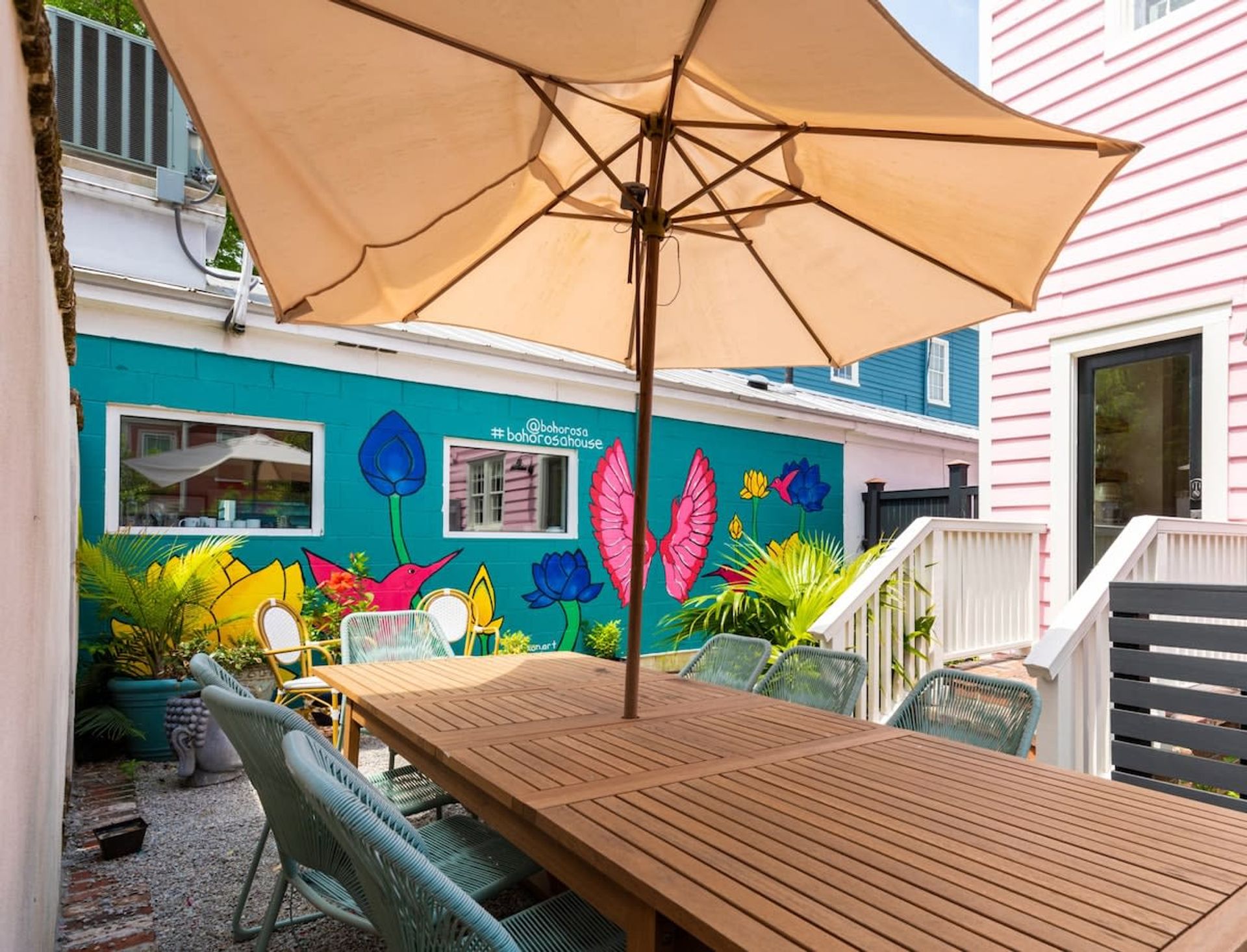 Vibrant courtyard patio with a long wooden dining table, large beige umbrella and woven chairs in front of a bright turquoise mural of flowers, birds and pink wings, flanked by pastel pink siding and potted tropical plants.