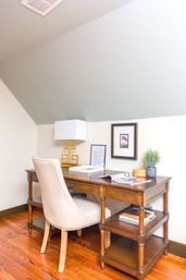 Cozy home office nook under a sloped ceiling with a wooden desk and beige upholstered chair, brass geometric lamp, framed artwork, potted plant and magazines on warm hardwood floors.