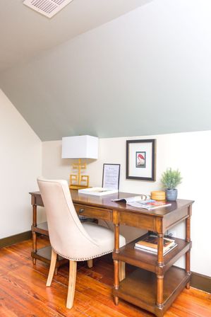 Cozy home office nook under a sloped ceiling with a wooden desk and beige upholstered chair, brass geometric lamp, framed artwork, potted plant and magazines on warm hardwood floors.