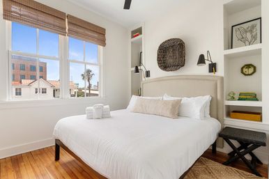 Bright beachy bedroom with crisp white linens, beige upholstered headboard, hardwood floors, woven window shades and built-in shelves, window view of palm trees and nearby coastal buildings.