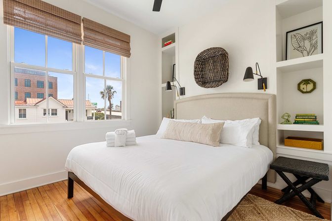 Bright beachy bedroom with crisp white linens, beige upholstered headboard, hardwood floors, woven window shades and built-in shelves, window view of palm trees and nearby coastal buildings.